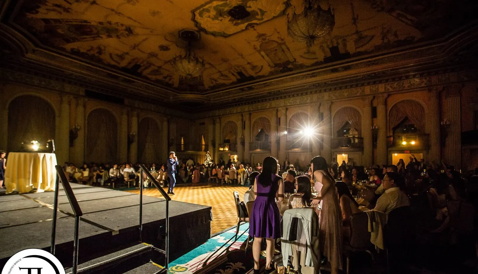 Johnny Wu performing on stage in a grand ballroom under a painted ceiling, with the audience seated at round tables