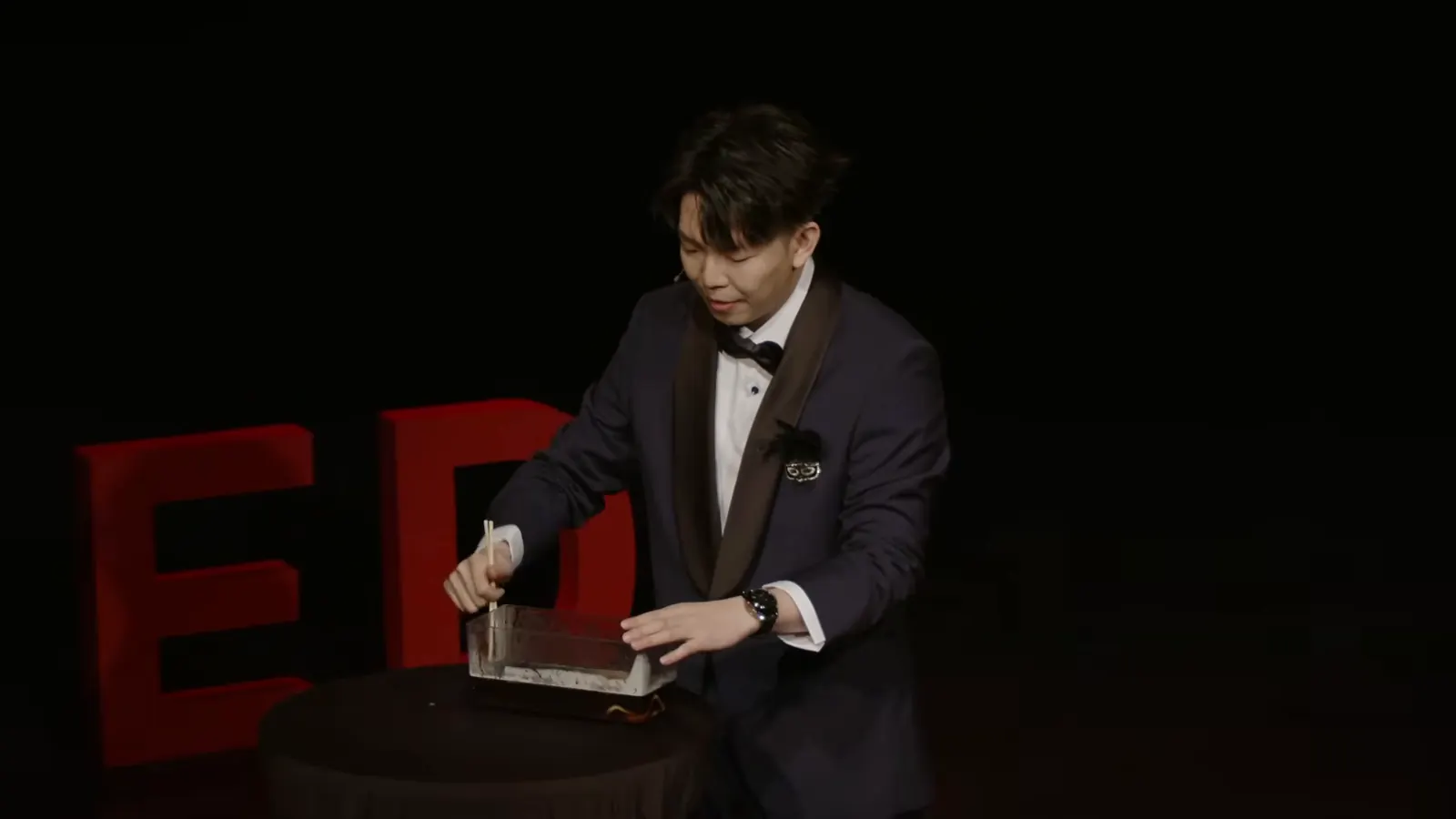 Johnny Wu at a table on the TEDxBeverlyGrove stage, in a tuxedo, with the red TEDx letters behind him